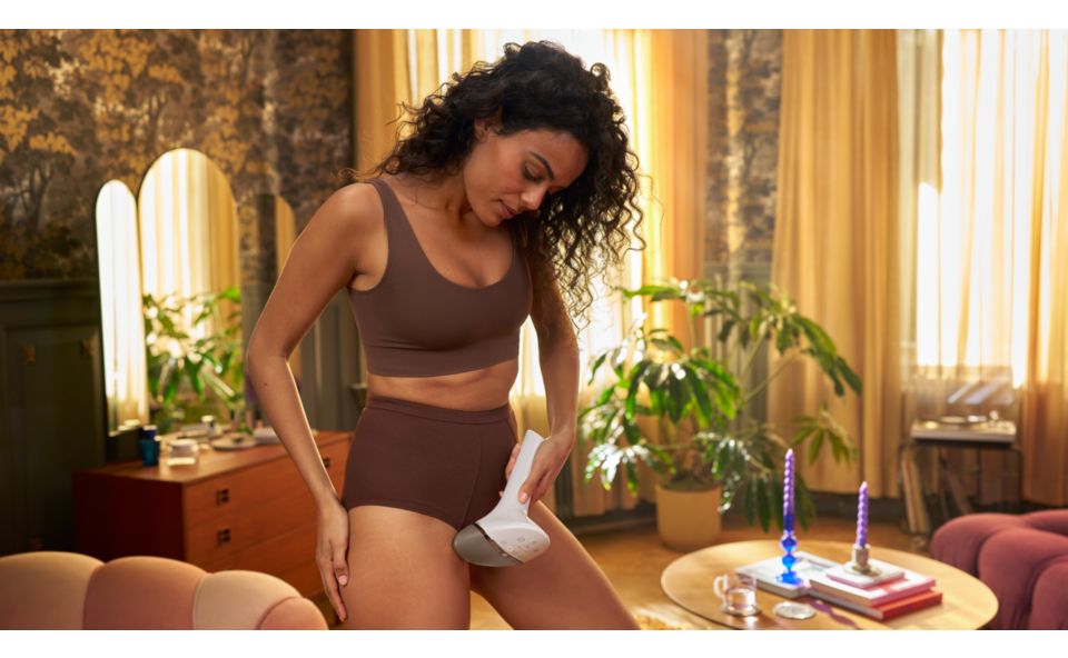 Kobieta używająca urządzenia IPL do depilacji okolic bikini, siedząca na fotelu w beżowym stroju kąpielowym.