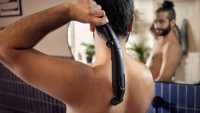 Un hombre utiliza una afeitadora corporal Philips para afeitarse el vello de la espalda mientras se mira en un espejo.