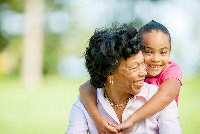 A smiling older woman is embraced from behind by a young girl, both enjoying time together outdoors.