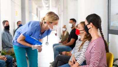 Nurse wearing a mask talking to a patient and child in a hospital waiting area during routine care