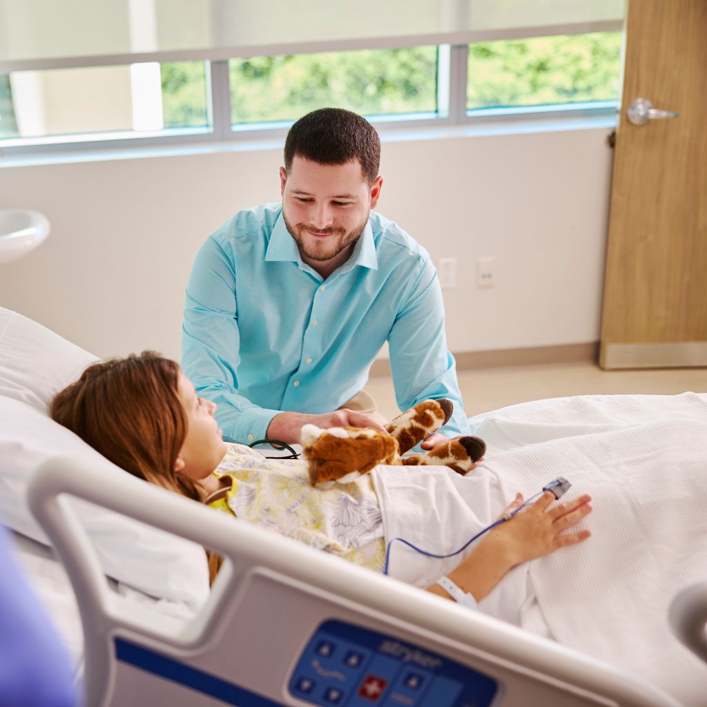 Pediatric patient in hospital bed with male caregiver and stuffed giraffe