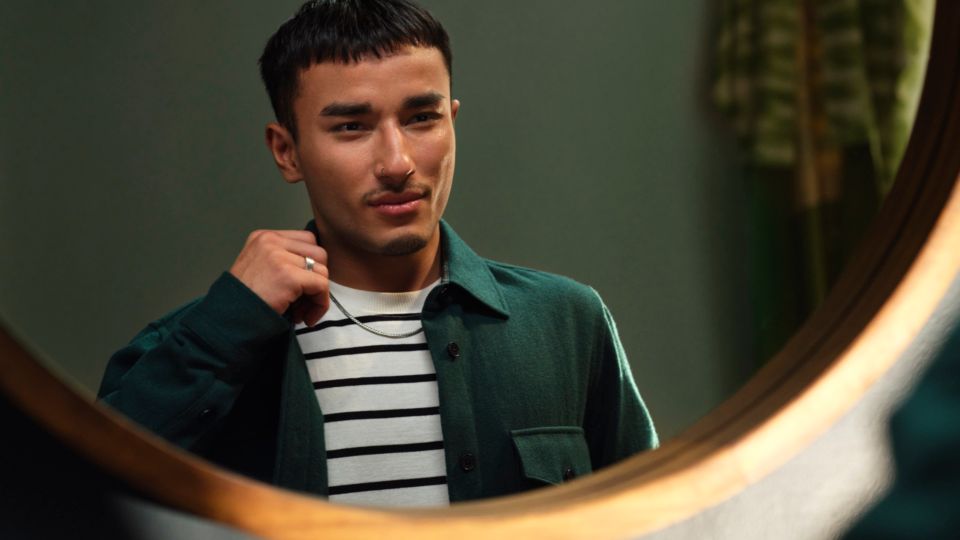 A young man with a thin moustache adjusts the collar of his green shirt in the mirror.