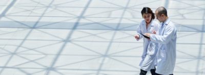 Two healthcare professionals in white lab coats walking together while reviewing information on a digital tablet, with geometric shadows cast across the bright floor.